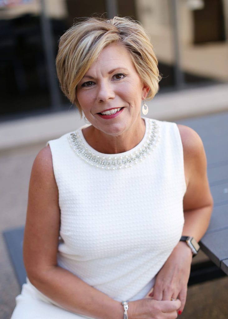 A woman with short blonde hair, wearing a sleeveless white textured dress with a beaded neckline, sits outdoors at a table, smiling at the camera. She has earrings, a bracelet, and a smartwatch.
