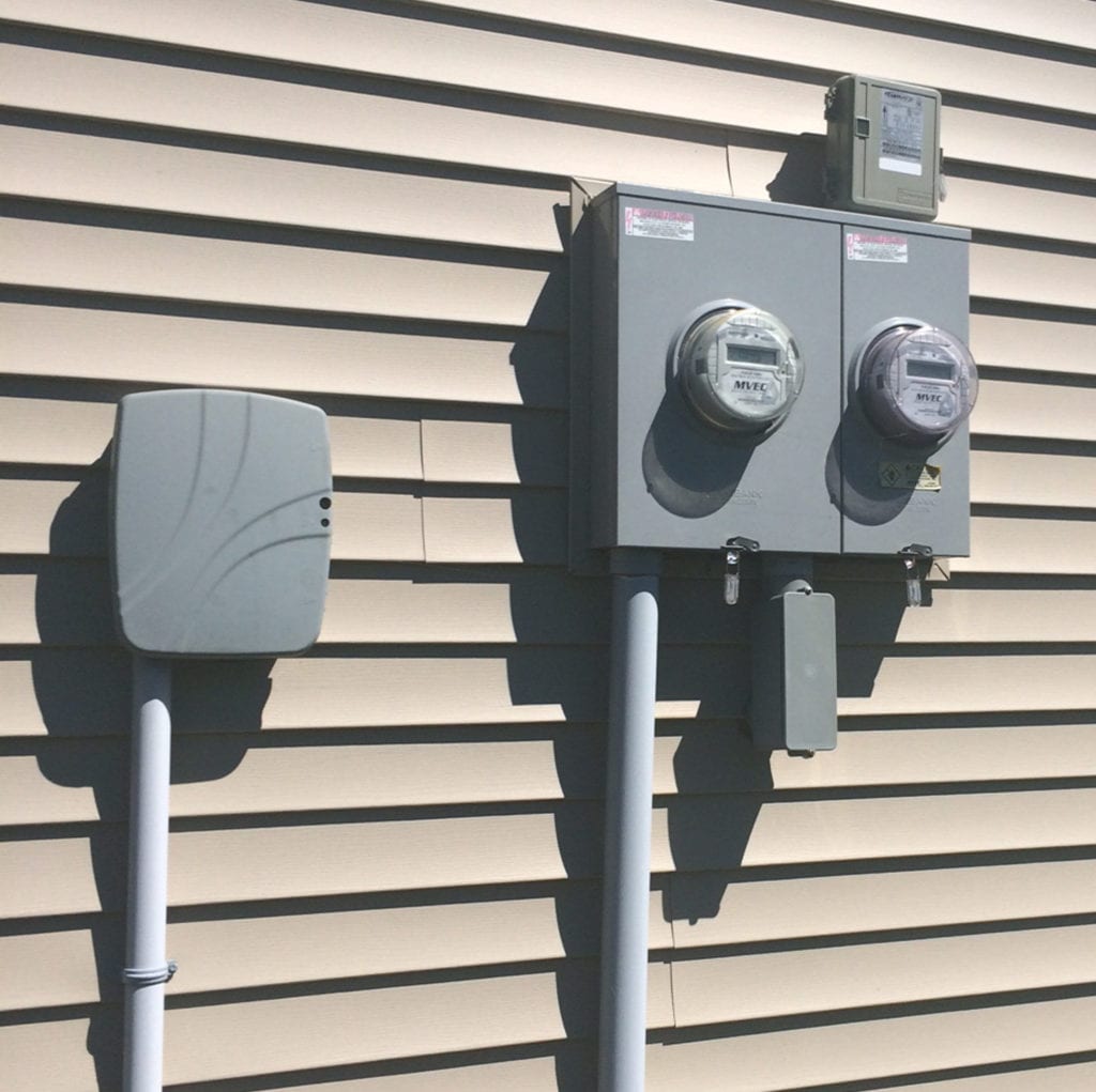Two electrical utility meters and a gray utility box are mounted on the beige vinyl siding of a building, with conduit pipes running vertically from each box to the ground.