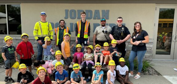 A group of children wearing yellow construction hats sit and stand in front of a building with JORDAN on the wall, posing with adults in safety vests, police uniforms, and casual clothes, smiling for the camera.