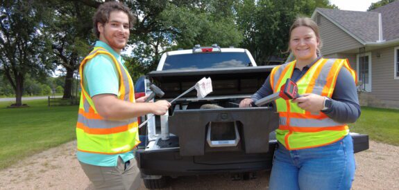 Two people wearing orange safety vests stand behind a pickup truck, holding litter grabbers and a trash bag. They are smiling and standing on a gravel driveway, with a house and trees in the background.