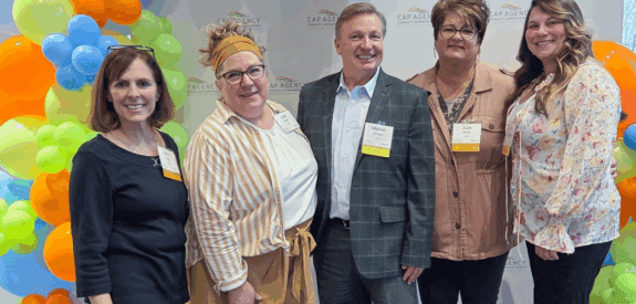 Five adults stand smiling in front of a CAP Agency backdrop, with colorful balloon decorations on the side. Three women are on the left, one man is in the center, and another woman stands on the right.