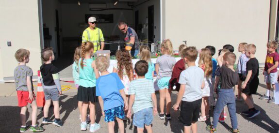 A group of children gather outside around two adults demonstrating something on a table in front of an open garage. The weather is sunny and the children appear attentive and curious.