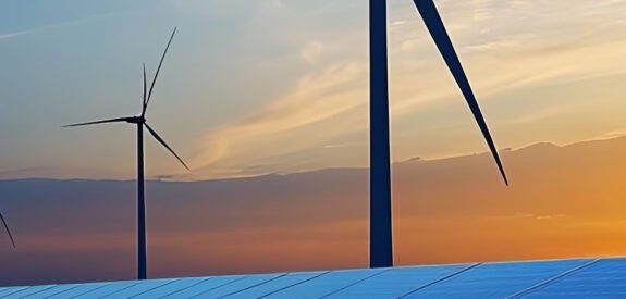 Wind turbines stand behind rows of solar panels at sunset, symbolizing renewable energy sources against a colorful sky.